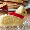 In a crystal dish, beside a plate of pancakes, butter formed in the shape of a lamb with a red ribbon around its neck, red flag sticking out from its back, and a black peppercorn for an eye.