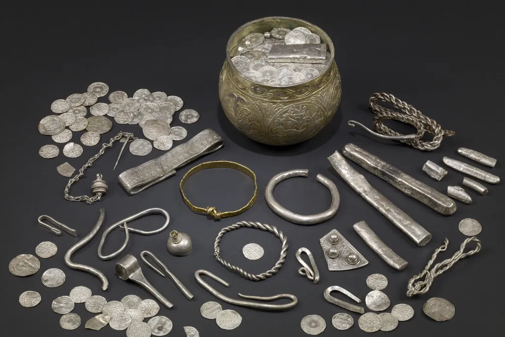 A display of silver Viking artifacts laid out on a table, including a wide mug-like cup, many coins, silver bars and what appear to be bracelets and other jewelry scattered around