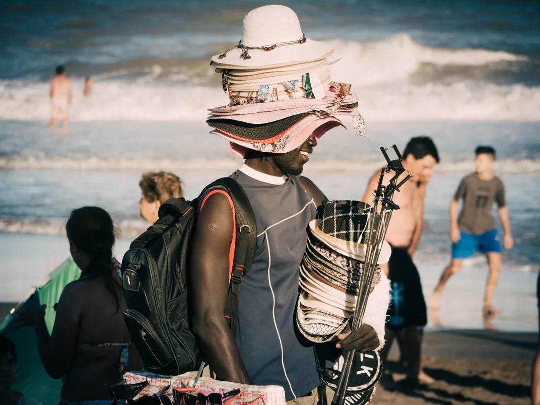 A Black man sells hats on the beach. He has several hats stacked on his head
