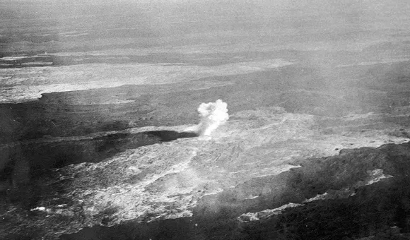 An old photo, tinted brown, depicts a flat barren landscape with a single pillar of dense white smoke at the center of the photo.
