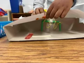 The hands of a student adjust the paper top of a playground model made with various crafting materials