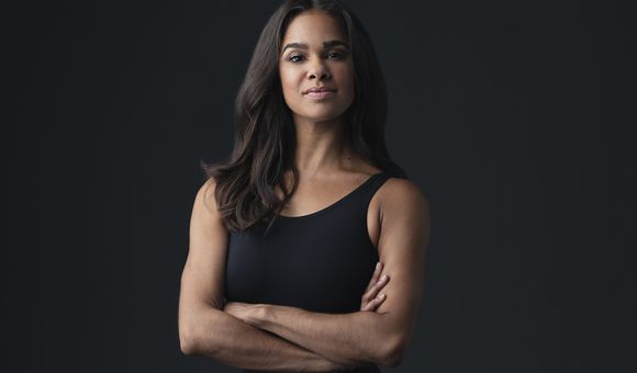 A woman stands with her arms crossed looking intently at the camera. She appears in front of a black background wearing a black leotard.