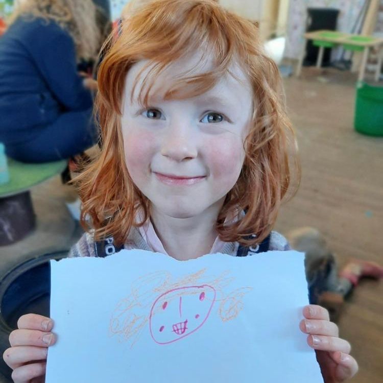 A pale, red-headed girl with bangs smiles and holds a piece of paper in front of herself, with what looks like a drawing of a person's smiling face and orange curly hair