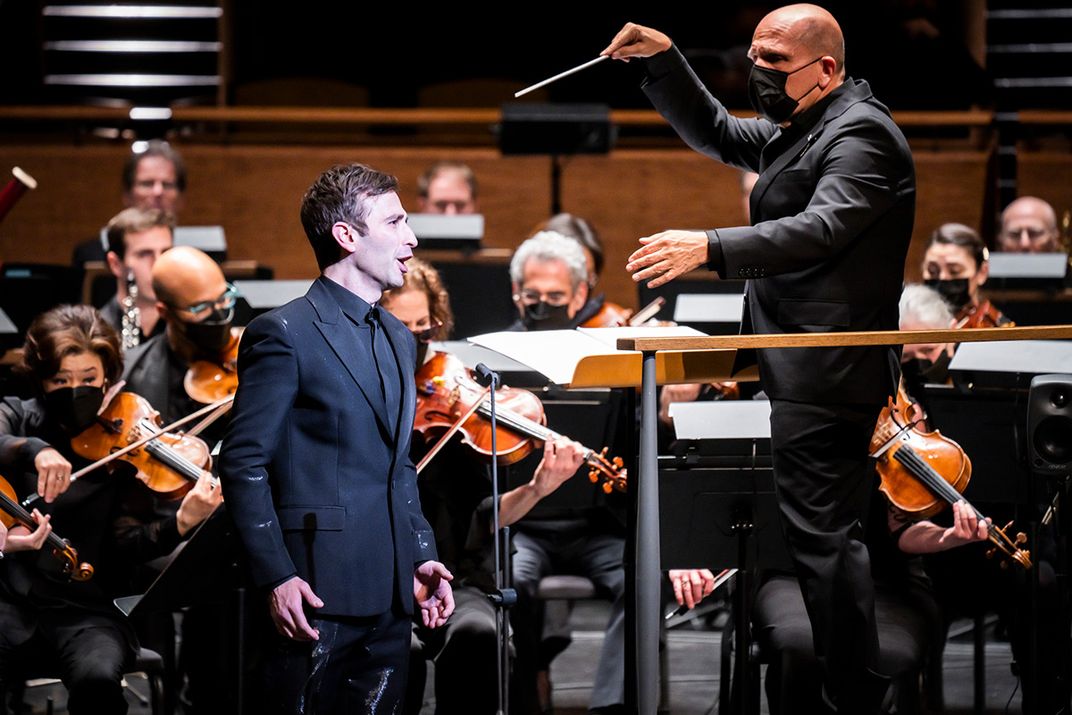 In the foreground, a man in all black suit sings, standing next to a conductor, also in all black clothes and face mask, on a podium. Behind them, members of an orchestra playing viola, violin, and woodwinds.