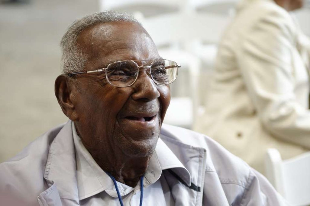 A close-up of Brooks, a black man with glasses and gray hair, smiling and wearing a white collared shirt and overcoat; he is sitting down and uses a wheelchair (not pictured)