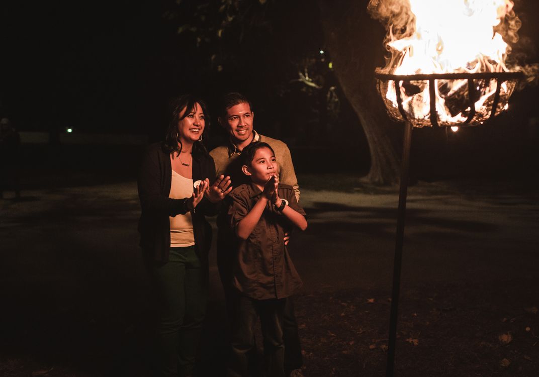 A group of three people stands outdoors at night beside a large flaming torch, illuminated by its warm glow against a dark wooded backdrop.