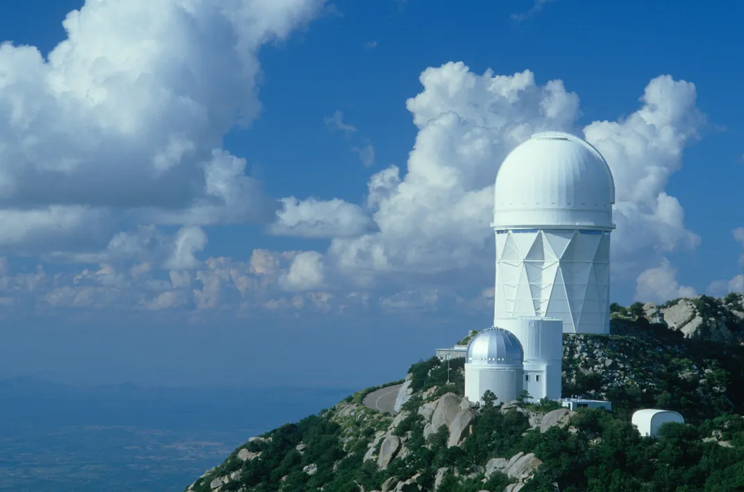 The Kitt Peak National Observatory sits on a hill with a cloudy blue sky in the background