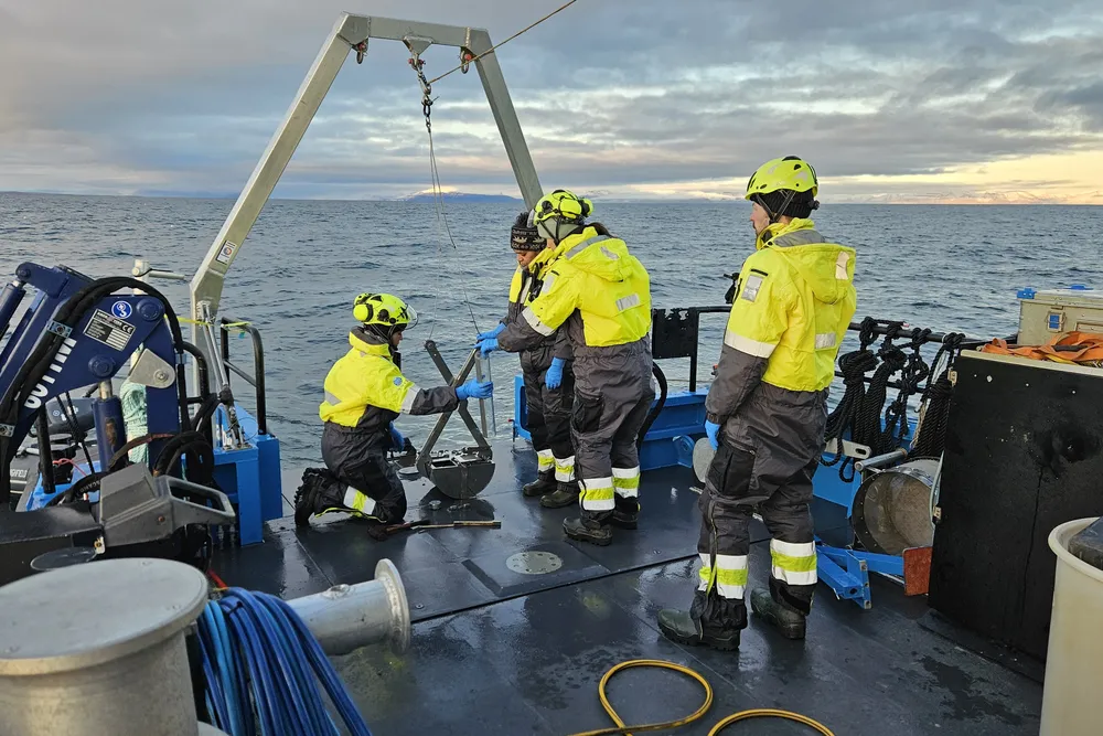 researchers in yellow hats and coats stand—one kneels—on the deck of a small research vessel on the ocean, examining a sediment core