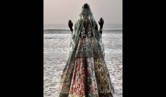 a person stands  in a decorated dress and vail on a beach with the sea behind them