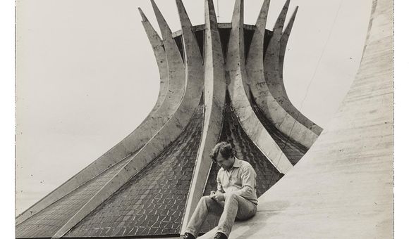 Grayscale image of Robert Hughes sitting on the sloping curve of a building examining something in his hands, with a modernist church in the background.