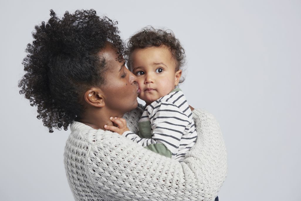 A mom holds her toddler and gives him a kiss on the cheek against a grey background