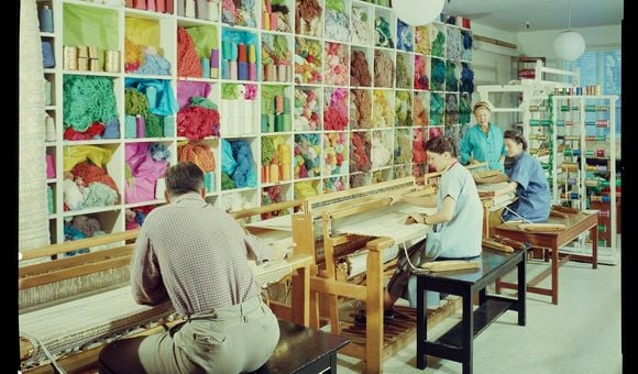 A smiling Dorothy Liebes stands in her studio in her blue apron, supervising two young women sitting at looms with spools of colorful thread and skeins of yarn close at hand.