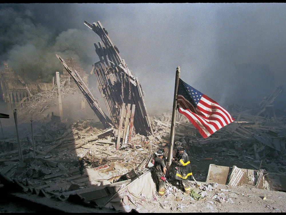 A dramatic horizontal view of the scene, with the shadow of a building cutting across the left side of the frame and the bright red stripes of the flag unfurling on its right, and an overwhelming amount of wreckage, dust and smoke in between
