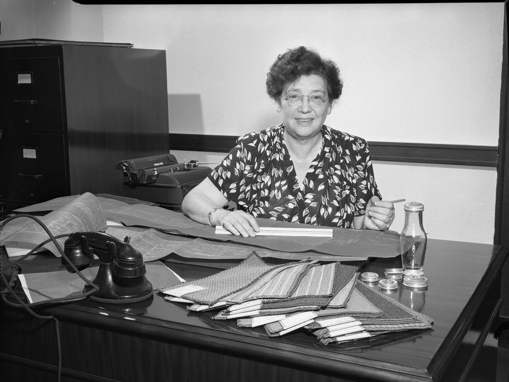 Black and white photograph of a woman sitting behind a desk. She has short, wavy hair and wears glasses. The desk is covered in rectangular carpet samples and blueprints are spread across the desk. A black telephone rests on the desk amid the supplies.