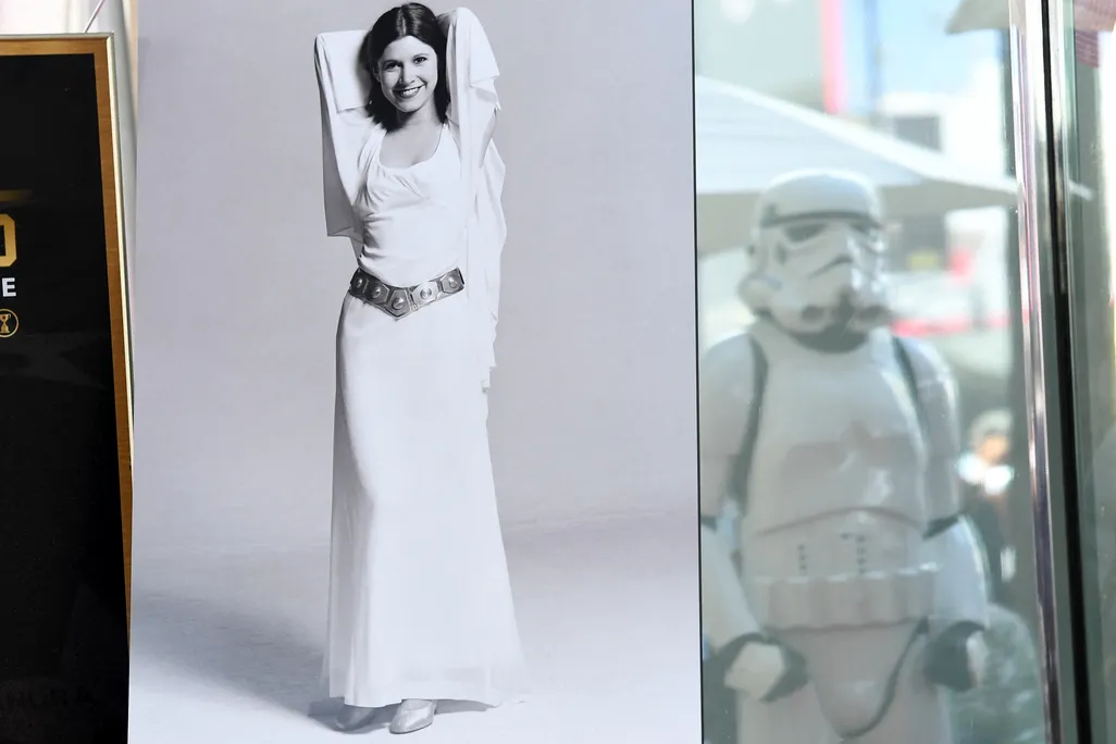 Photo of Carrie Fisher wearing white gown, with white storm trooper in background