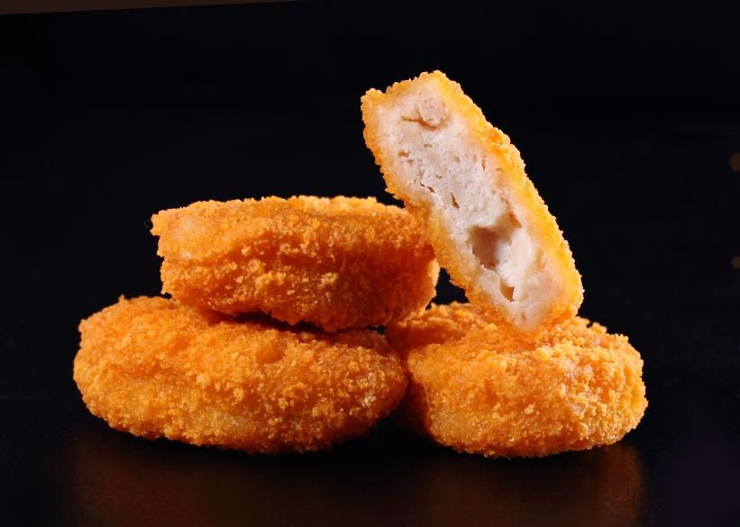 A close-up shot of three chicken nuggets stacked up in front of a black background. On top is one chicken nugget, cut in half, with the insides facing the camera. 