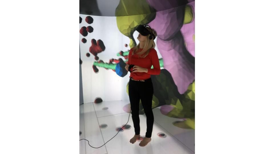 Dr. Arcand is standing viewing a wall of red, green, blue, yellow, and pink 3D visualization through a black headset that covers her eyes and ears.  She is wearing a red long-sleeve shirt, black pants, and is not wearing shoes or socks.