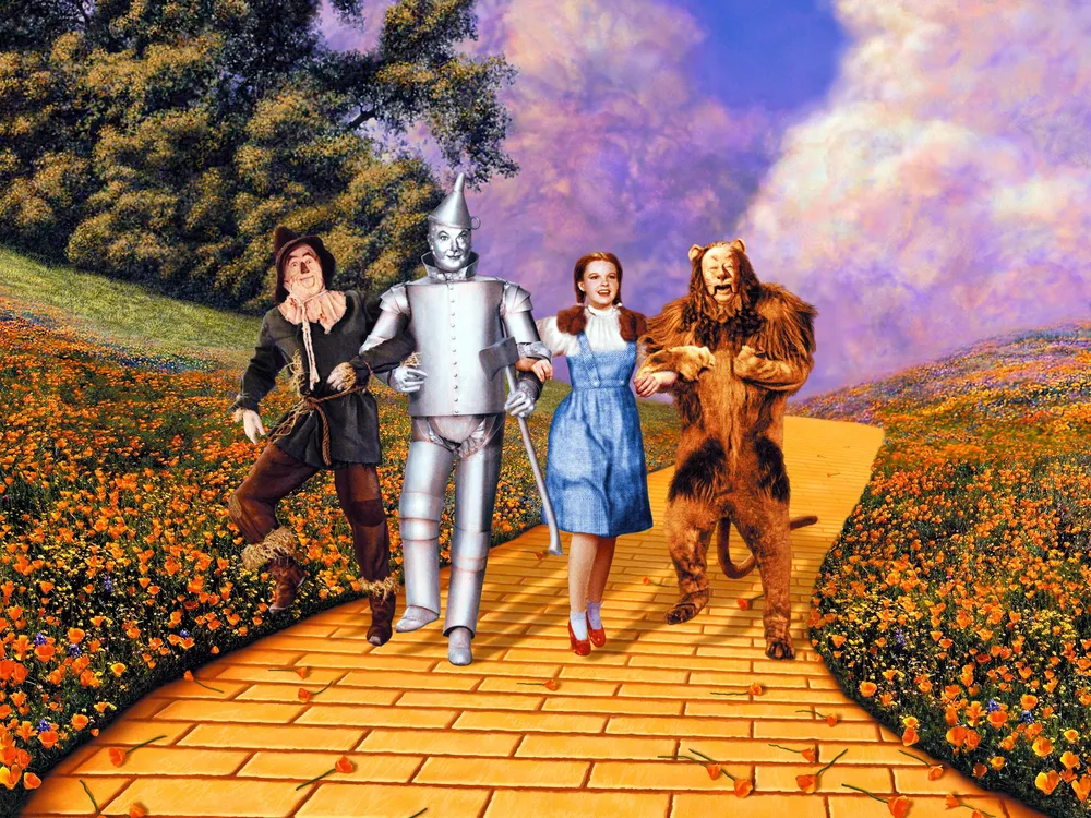 Four people, a scarecrow, Tin Man, Dorothy and a lion, walk arm in arm down a yellow brick road 