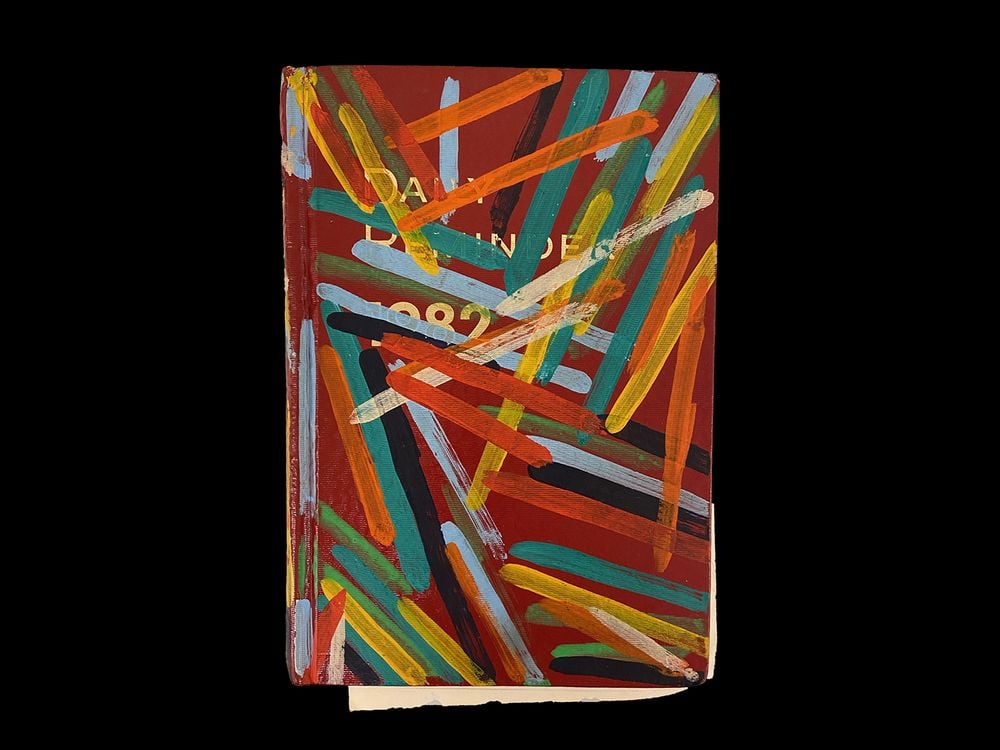 A photograph of a red book that reads, “Daily Reminders 1982” in white lettering. The book has yellow, green, blue, red, and orange strips of paint on it.