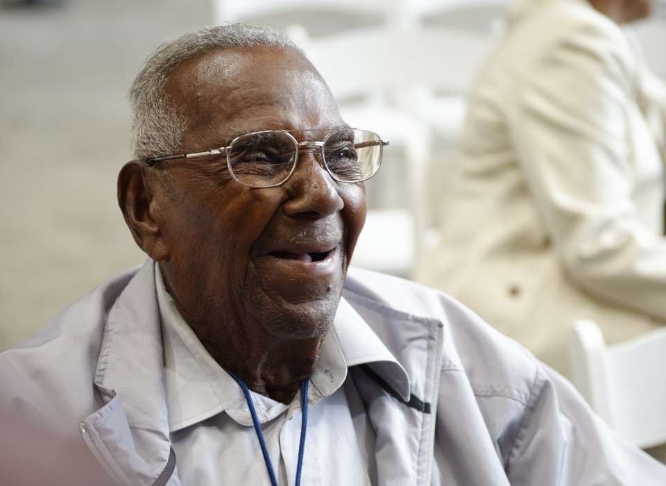 A close-up of Brooks, a black man with glasses and gray hair, smiling and wearing a white collared shirt and overcoat; he is sitting down and uses a wheelchair (not pictured)