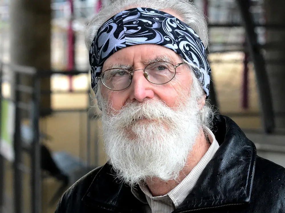 A close-up Michel Talagrand, who wears a black leather jacket, glasses, and a blue bandana wrapped around his forehead, looking to the right of the camera.