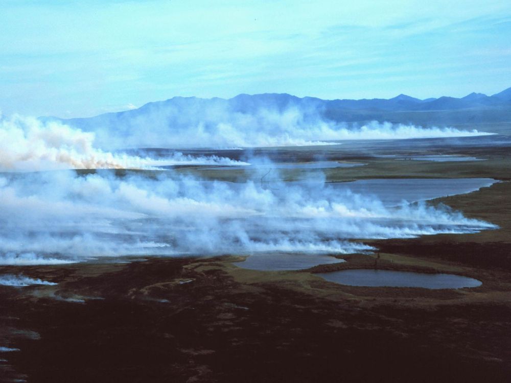 A photograph of a tundra landscape with mountains on the horizon and smoke rising from the ground