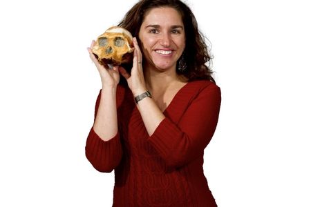 Headshot of paleoanthropologist Briana Pobiner in a red sweater holding an early human skull up text to her face on white background.