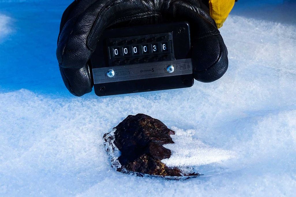 A close-up photograph of a black meteorite half-submerged in white snow and ice. A black and yellow gloved hand holds a small measuring device next to the rock.