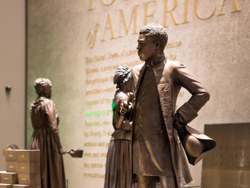 A bronze colored statue of Bannecker, a Black man wearing a suit jacket and high boots, stands in a museum exhibition