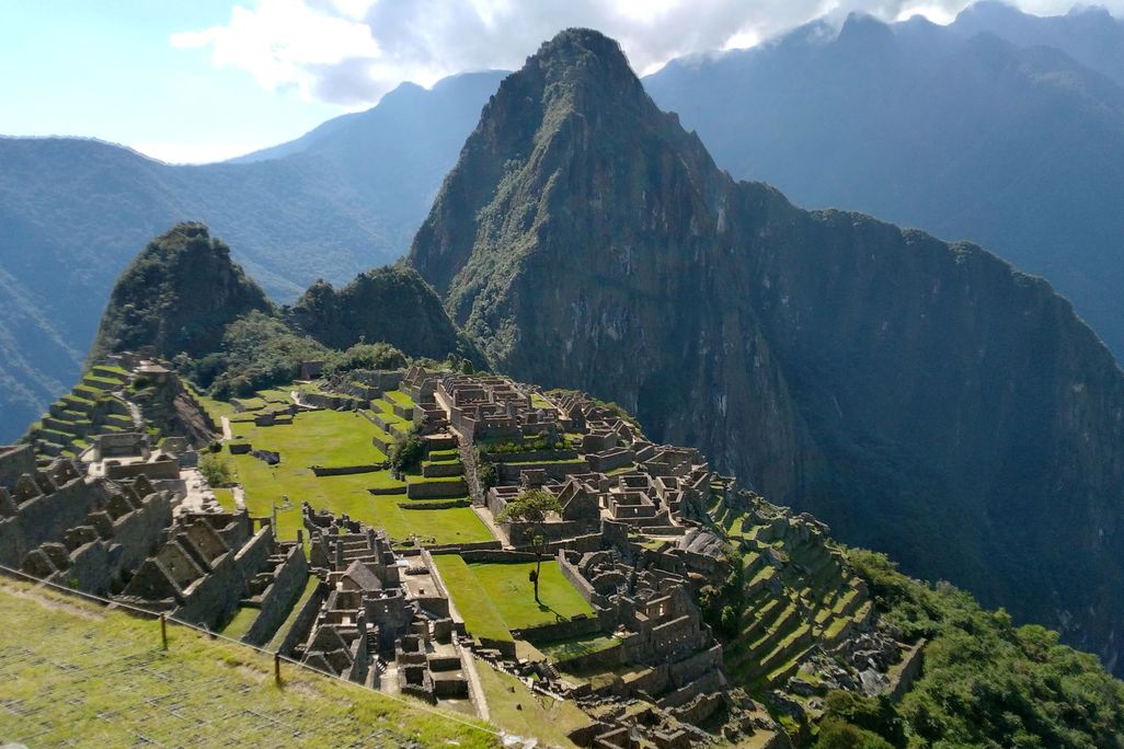 A panoramic view of Machu Picchu from above, with green slopes and angular mountain ridge in distance and the ruins of settlements along a sloping green mountainside; devoid of tourists