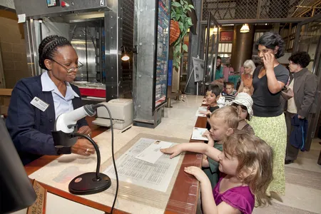 A group of students wait in line at a Post Office. Each student has a stamped envelope they are handing to the postal worker behind the counter.