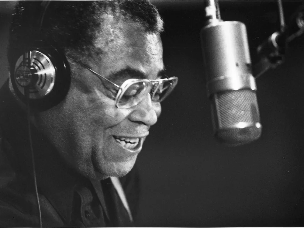 Black and white photo of a Black man wearing headphones and speaking into a microphone