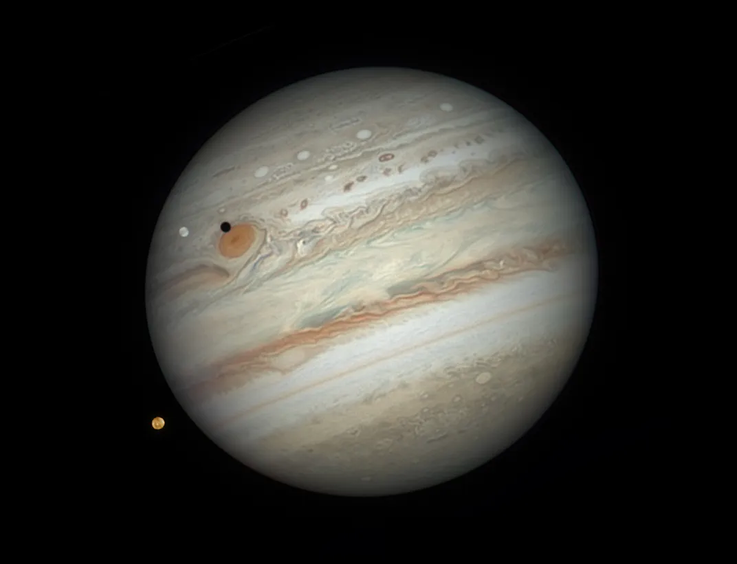 jupiter, with the red spot on its upper left, and two of its moons; europa appears white above the planet's surface, and Io appears orange at the lower left