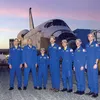 Seven people wearing blue flightsuits stand in front of a space shuttle parked outside.