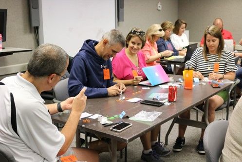 A group of educators sit around a rectangular table with various design materials, phones and laptops.