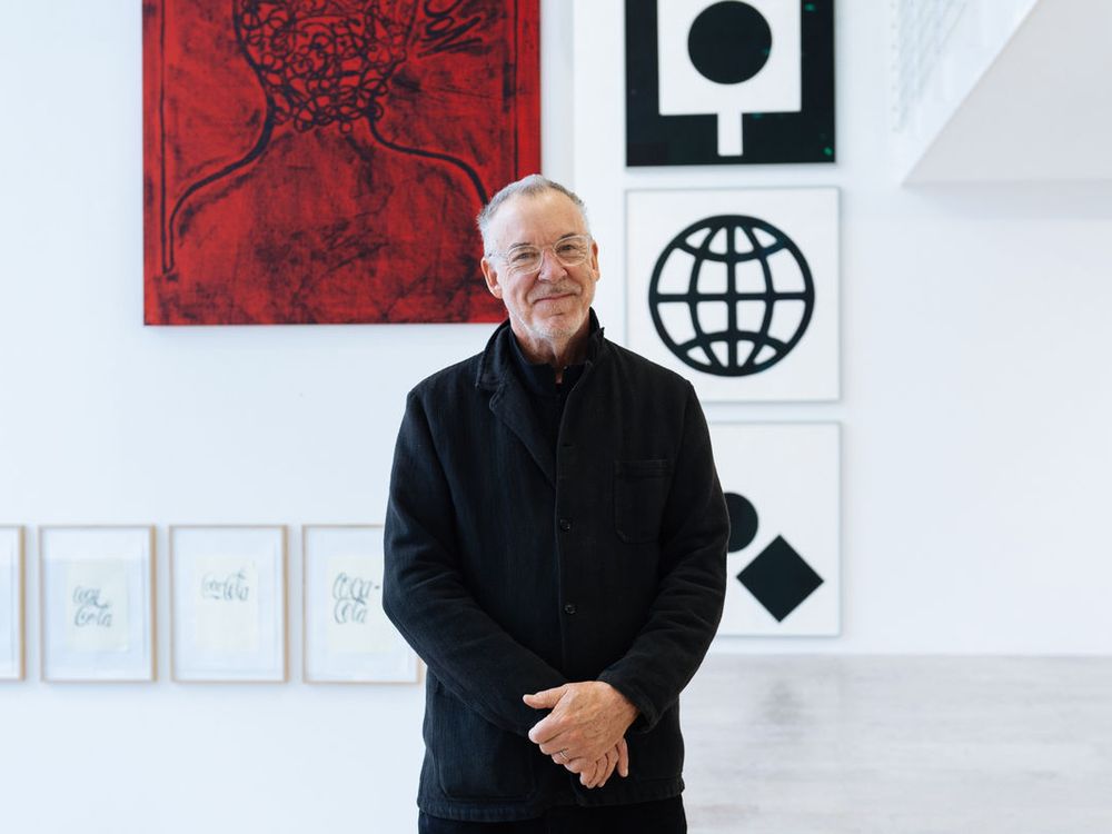 Artist Matt Mullican wearing glasses and a black jacket with his hands folded in front of his body, standing in front of six artworks, with his body partially hiding them.
