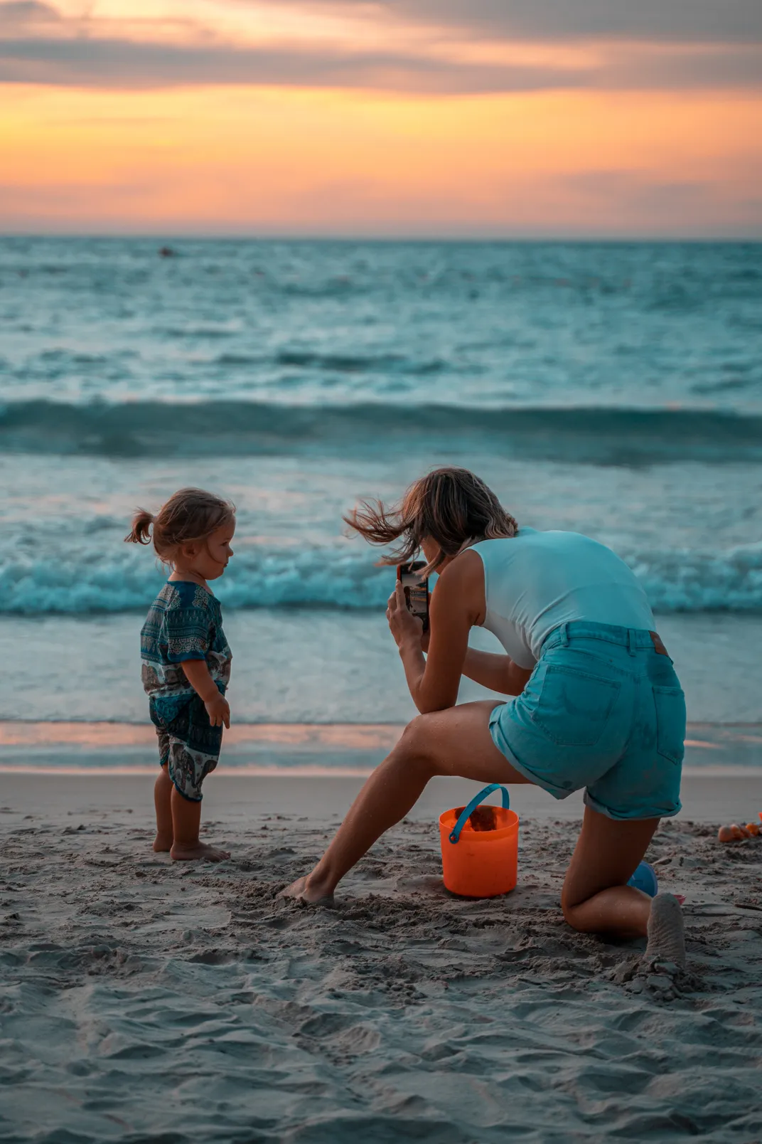A mother snaps a photo of her young daughter on the beach at sunset.