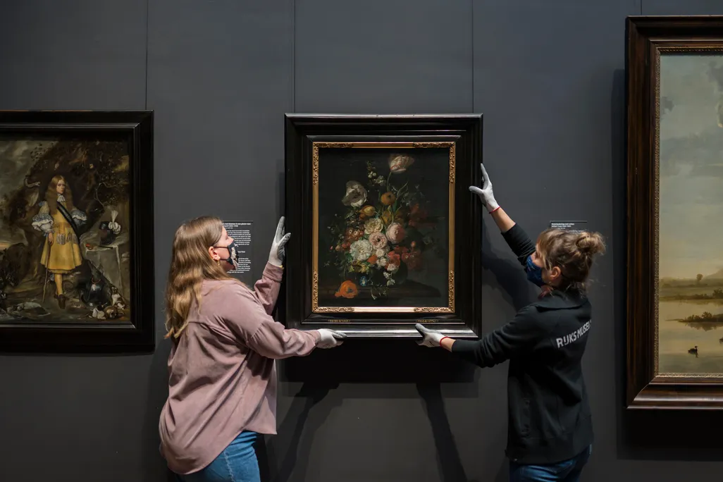 Two people in gloves and masks hold either side of the painting, which depicts a lush bouquet, and hangs it on the dark gallery wall