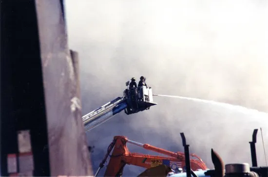 Photo of World Trade Center site after attacks of September 11, 2001. Two or three workers in hard hats and uniforms are in the cherrypicker of a crane, shooting a fire hose downward in a gentle arch. A battered wall or part of a building visible left.