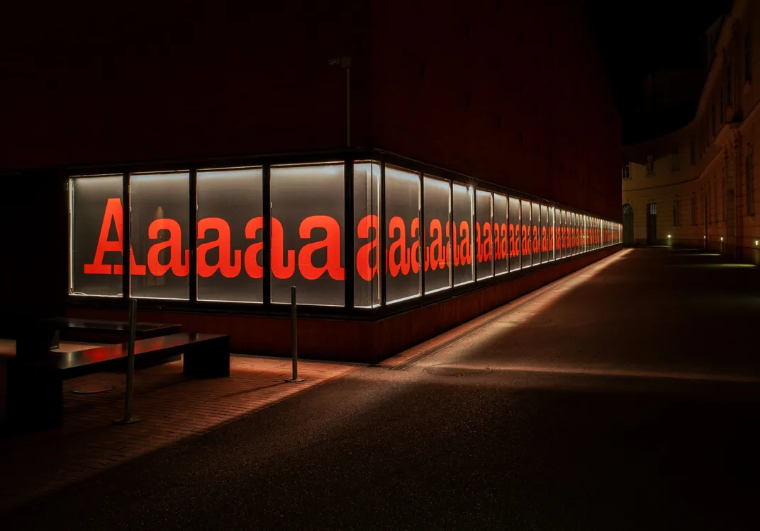 Night view of a modern glass building displaying large red letters "A" and "a" in its windows, illuminated from within.