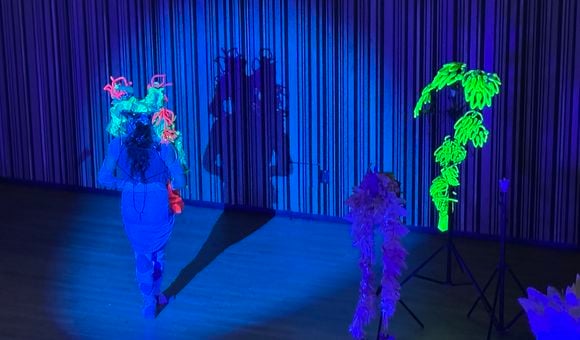 Armando Lopez-Bircann wears a cobalt blue bodysuit and skirt on a stage bathed in blue light. They have a neon green, blue, and pink headdress on and face away from the audience, with in front of their shadow. To the right are props including a neon green