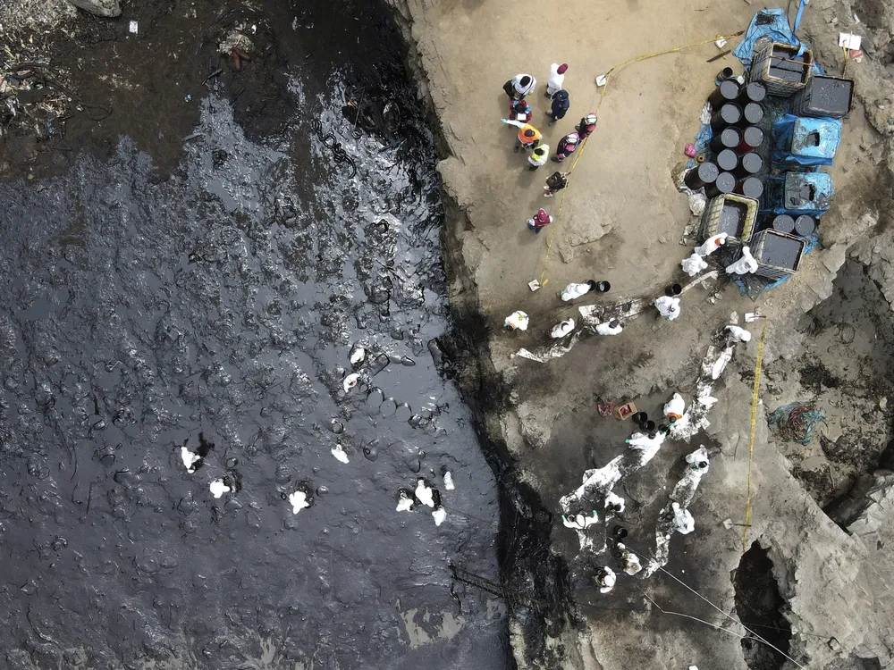 An aerial image of an oil spill off Peru's coast. The photo shows a contaminated beach smeared with black crude oil and workers in PPE trying to clean up the beach.