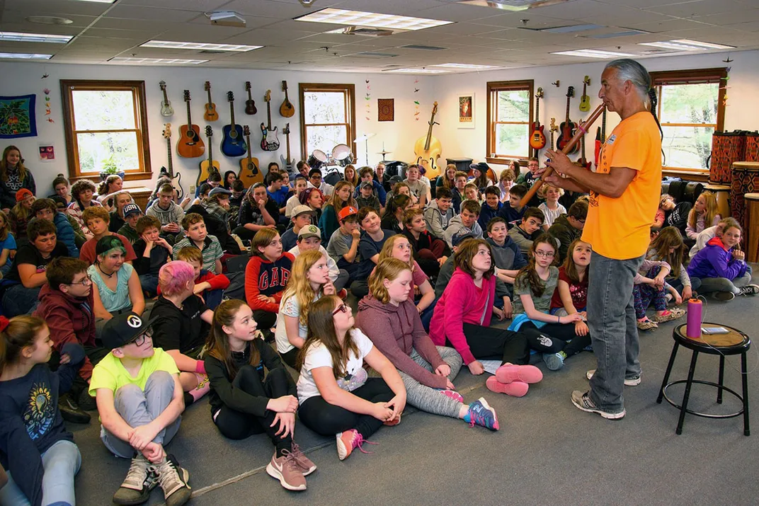 Man with long braids and bright yellow shirt plays flute in front of a packed classroom of kids sitting on the floor.