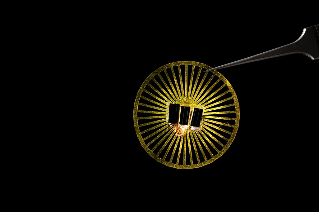An image of a small wheel like yellow disc with sensors attatched to the middle. A set of tweezers is holding the device against a black background.