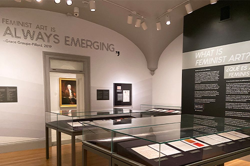 An image of the 'What is Feminist Art' exhibition with wall quote reading 'Feminist art is always emerging' and a portrait of a man seen through the doorway