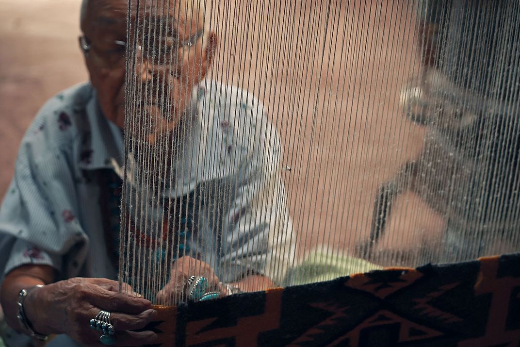 An elder woman weaves on an upright loom, sitting on the ground.