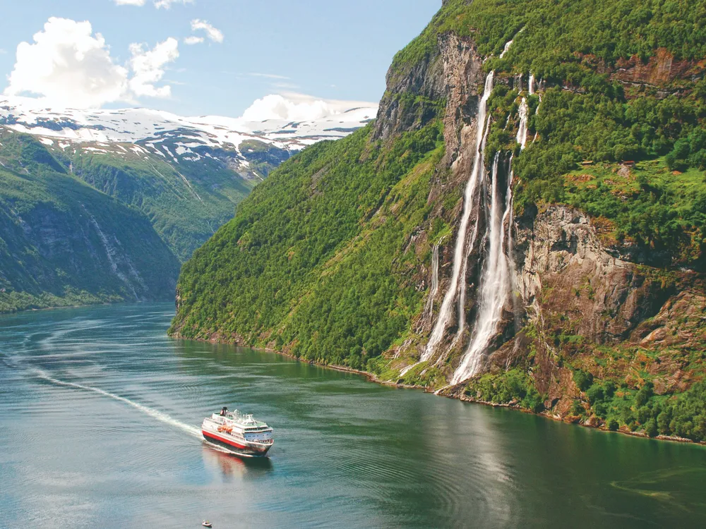 A cruise ship sails through Geirangerfjord in Norway, surrounded by steep green mountains, dramatic waterfalls cascading down rocky cliffs, and distant snow-covered peaks beneath a bright blue sky.