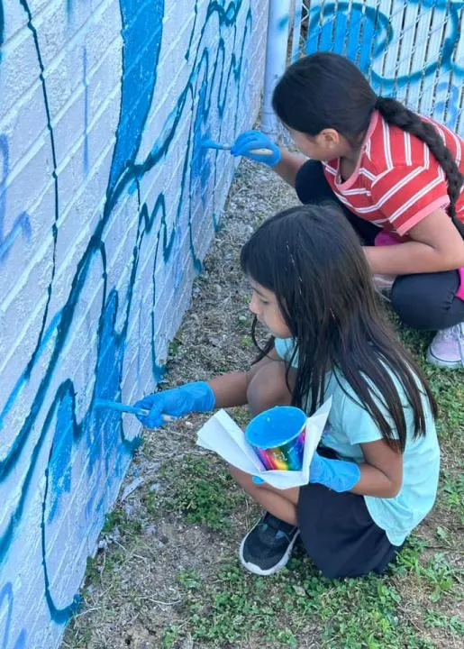 Two kids wearing blue latex gloves crouch painting blue shapes on a brick wall.