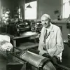 Black-and-white image of a white man in his early 50s. He is bald and has a mustache. Wearing a white lab coat, he is sitting at a work table supporting a large metal cylinder.