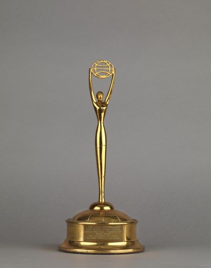 A gold award. There is a circular base with engravings on the front and a slim figure holding up a flat circle with lines running across it. 